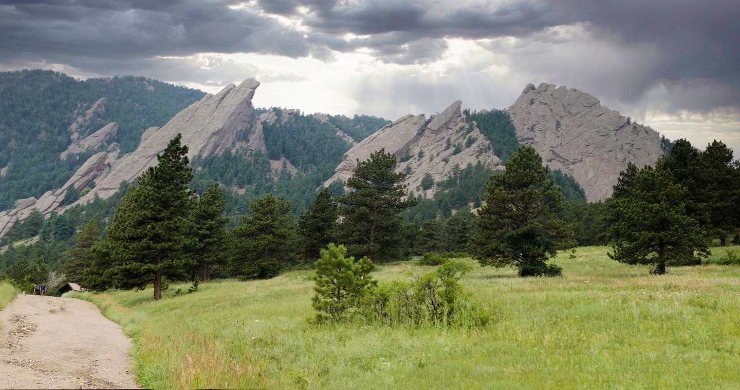 The Flatirons rising above a summer meadow and pines on Bluebell Road, Boulder County, Colorado, under broken storm light